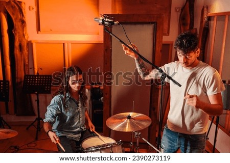 Picture of a sound technician assisting with setting up recording microphone for the drummer. The drummer is playing drums, long hair and stylish glasses. A recording studio worker is adjusting a mic