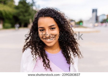 Happy young latin american girl smiling at camera standing at city street. Outside portrait of joyful beautiful woman over urban background. Royalty-Free Stock Photo #2390283837
