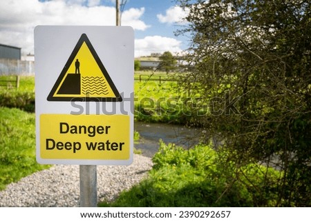 Yellow, black Danger Deep Water sign close up on riverbank of rural waterway river stream. Green fields in background. Hazard signage alerting public to dangerous deep water nearby. Drowning risk.