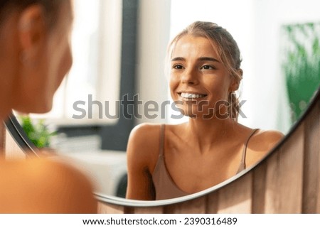 Smiling beautiful confident woman with perfect makeup wearing tank top looking in mirror standing at home. Natural beauty, skin care, morning routine concept Royalty-Free Stock Photo #2390316489