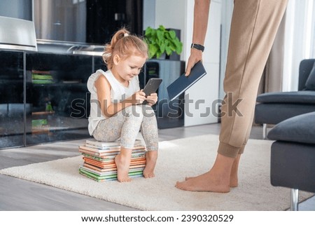 Little girl sits on a stack of children's books and plays with her smartphone while mother gives her a book. High quality photo
