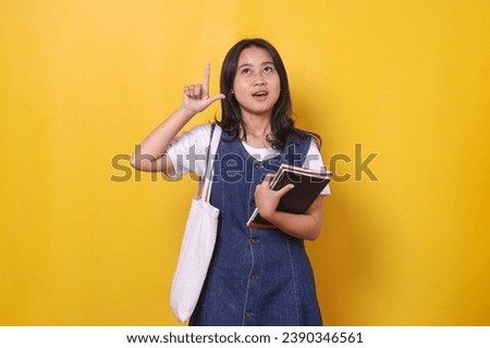 Young Asian student girl holding book and pointing above for educational ads