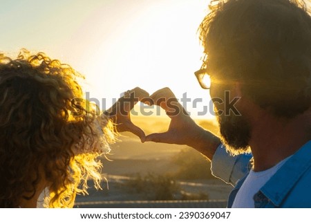 Romantic couple in love doing heart symbol with hands against an amazing golden sunset. Man and woman flirting and dating outdoor enjoying nature. People and life together forever concept. Travel day