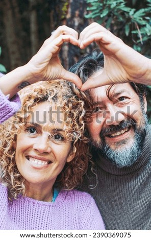 Happy adult couple people in love do hearth symbol gesture with hands together smiling and enjoying relationship. Man and woman leisure with green foliage woods forest in background.