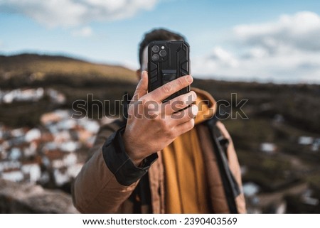 Young happy photographer man traveling sightseeing in a small town, taking a photo with his cell phone of the panoramic view of the town and the mountains, from a castle on a sunny day