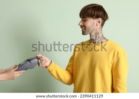 Tattooed young man paying with credit card via terminal on green background