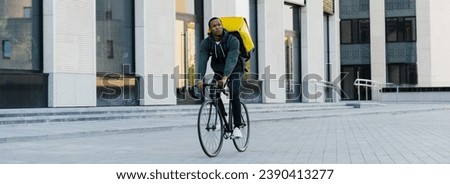 An African-American deliveryman with a yellow backpack rides a bicycle against the background of a modern business center. Panoramic photo.