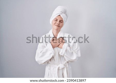 Blonde caucasian woman wearing bathrobe smiling with hands on chest with closed eyes and grateful gesture on face. health concept. 
