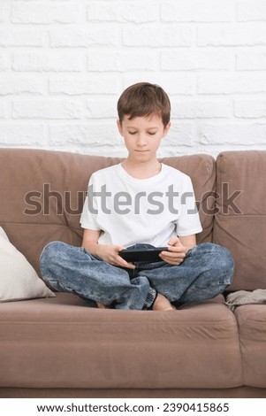 Close up cute little boy wearing headphone using smartphone, looking at screen, child holding phone in hands, sitting on couch at home alone, playing mobile device game, watching cartoons online.