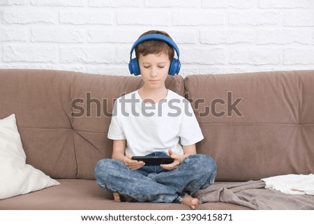 Close up cute little boy wearing headphone using smartphone, looking at screen, child holding phone in hands, sitting on couch at home alone, playing mobile device game, watching cartoons online.