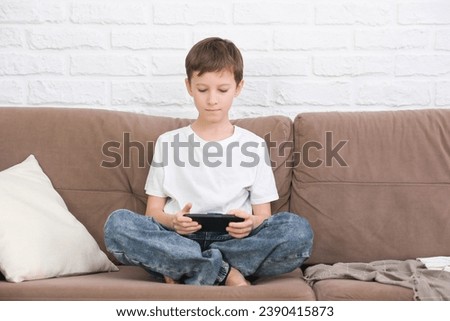 Close up cute little boy using smartphone, looking at screen, child holding phone in hands, sitting on couch at home alone, playing mobile device game, watching cartoons online.