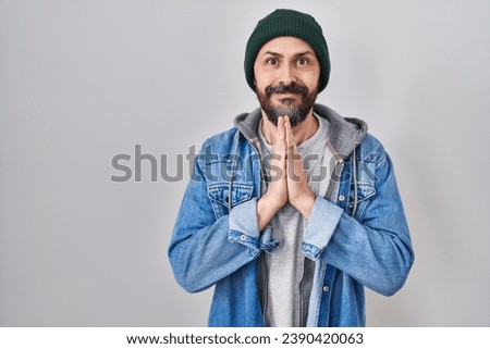 Young hispanic man with tattoos wearing wool cap praying with hands together asking for forgiveness smiling confident. 