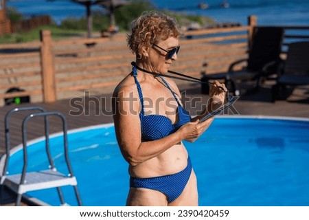 An elderly woman in sunglasses and a blue bikini is sunbathing by the pool. The pensioner uses a water-resistant phone case.