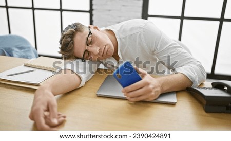 Exhausted young caucasian businessman, smartphone in hand, loses battle with stressâ€“ nobly crashes asleep on office table, job tedium takes its toll. Royalty-Free Stock Photo #2390424891