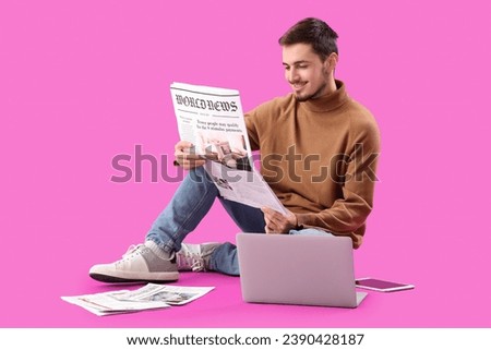 Young man with newspaper and laptop sitting on purple background