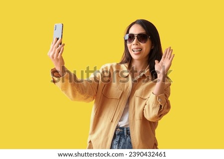 Beautiful young Asian woman with mobile phone waving hand on yellow background