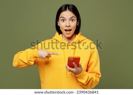 Young surprised shocked Latin woman she wears yellow hoody casual clothes hold in hand use point index finger on mobile cell phone look camera isolated on plain pastel green background studio portrait