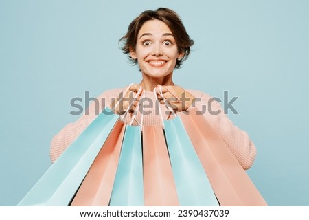 Young surprised woman wear beige knitted sweater casual clothes hold in hand paper package bags after shopping isolated on plain light pastel blue cyan background. Black Friday sale buy day concept