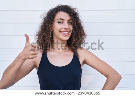 Hispanic woman with curly hair standing over white background smiling happy and positive, thumb up doing excellent and approval sign 
