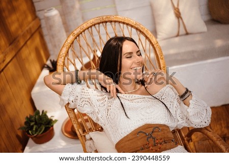 Joyful woman in knitted dress relaxing in wicker chair in modern bungalow	