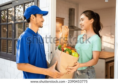 Asian young delivery man delivering package to female customer at home. Attractive postman in blue uniform working, deliver a box of fruit and vegetable groceries to woman infront of the door in house Royalty-Free Stock Photo #2390453775