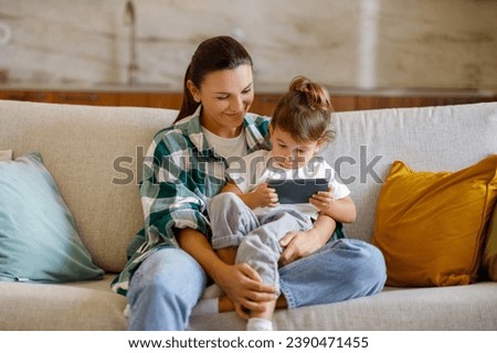Young Mother And Her Toddler Daughter Using Smartphone Together At Home, Mommy And Cute Little Child Watching Cartoons Online While Sitting On Couch In Living Room Interior, Copy Space