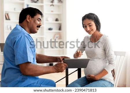 Smiling mature doctor showing digital tablet with test results to pregnant indian woman in clinic, male gynecologist in uniform consulting young expectant mother in modern hospital, giving advices Royalty-Free Stock Photo #2390471813