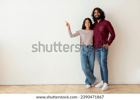 Happy positive beautiful loving young indian couple buying new house, pointing at blank copy space and smiling at camera at empty room in apartment, full length. Mortgage for millennials, rent