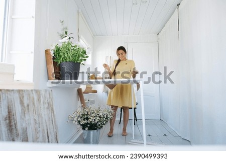 A barefoot woman in a yellow dress paints at a high table in a white home studio beautifully decorated with flowers,