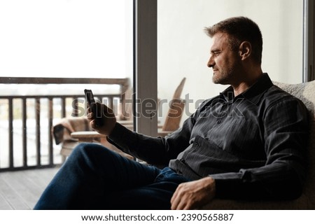 Caucasian middle age man using smartphone for planning his day. Confident male browsing social media when sitting on armchair. Relaxed father check photos of his family