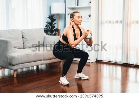 Athletic and sporty senior woman engaging in leg day training session with squat and bodyweight kettle ball at home exercise as concept of healthy fit body lifestyle after retirement. Clout Royalty-Free Stock Photo #2390574395