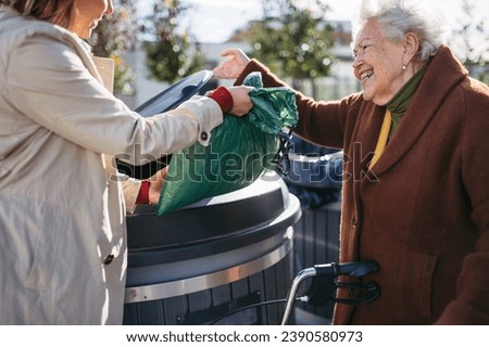 Woman helping elderly neighbor throw away trash into garbage can, waste container in front of their apartment complex. Royalty-Free Stock Photo #2390580973