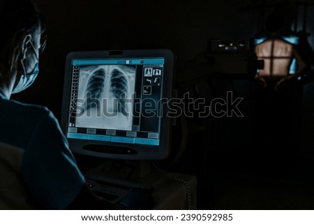Man standing face against wall while doctor using X-Ray machine scan him in darkness. Lungs x-ray image. Pulmonology, coronavirus prevention. Royalty-Free Stock Photo #2390592985