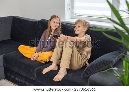 Children sitting on the sofa and watching TV interesting film. Boy holding TV remote. Rest after school. Royalty-Free Stock Photo #2390616361