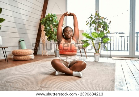Flexible body. Plump black woman doing stretching exercises for arms during morning exercise at living room. Young female in sport bra and shorts sitting in lotus pose and keeping eyes closed. Royalty-Free Stock Photo #2390634213