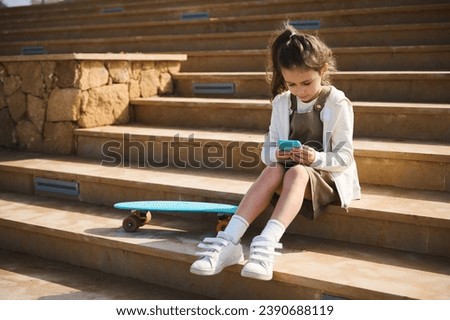 Adorable little child girl, sitting on steps near her penny board outdoors, using mobile phone, scrolling newsfeed, browsing internet, playing online video games, watching cartoons.