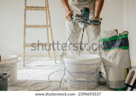 Builder's hands using manual cement mixer for mixing plastering materials and preparing it for skim coating and plastering walls in a house in renovation process. Cropped picture of man mixing plaster