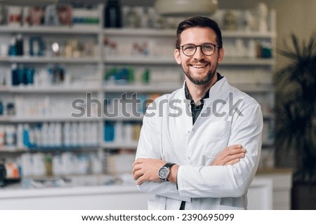 A happy young Caucasian pharmacy shop owner, standing in their newly-opened pharmacy shop. Wearing a white coat, glasses and a watch. The stature reflects success and high hopes for their business. Royalty-Free Stock Photo #2390695099