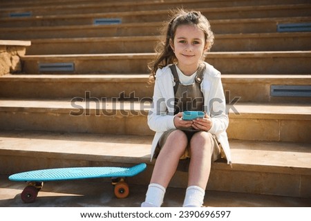 Authentic lifestyle portrait of a lovely little girl, elementary school student in casual wear, holding mobile phone, sitting on steps with her skateboard, smiles cutely looking confidently at camera