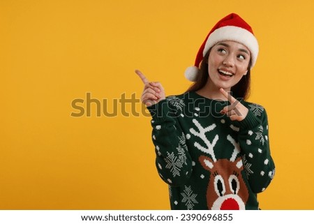 Happy young woman in Christmas sweater and Santa hat pointing at something on orange background. Space for text