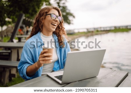 Portrait of a beautiful female freelancer working online using a laptop and enjoying the natural landscape overlooking the lake. Freelance concept, nature. Royalty-Free Stock Photo #2390705311