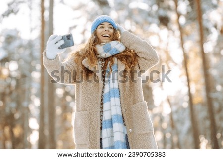 Happy woman in a bright hat and scarf with a phone in her hands takes a selfie, blogs in a sunny winter snowy forest. Travel concept, technology, fun.