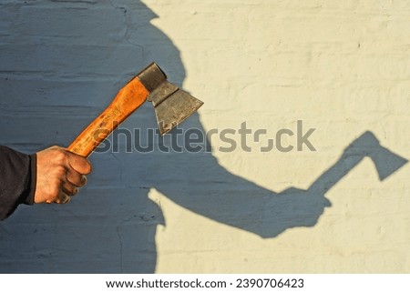 small old sharp dangerous iron industrial rusty with a wooden handle iron hatchet in hand near a white wall on the street with the shadow of a man's silhouette in the sun Royalty-Free Stock Photo #2390706423