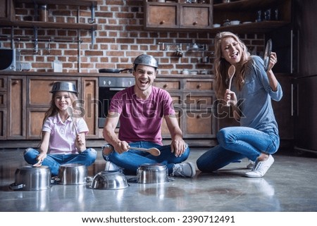 Cute little girl and her young parents are using wooden spoons and smiling while playing drums with dishes in kitchen Royalty-Free Stock Photo #2390712491