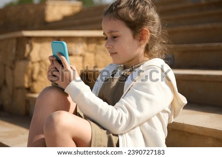 Portrait of a beautiful pensive happy elementary age Caucasian little girl with a thoughtful expression, sitting on the steps outdoors, engrossed in her smartphone, watching cartoons, online chatting