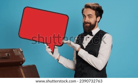 Hotel concierge shows ad with speech bubble, indicating empty isolated red cardboard icon. Skilled bellboy presents blank copyspace billboard sign in studio against blue background.
