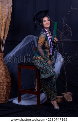 Beautiful exotic Asian woman graduating smiling happily holding diploma and thighs aside wearing green kebaya and elegant toga strap in indoor photo studio with black background