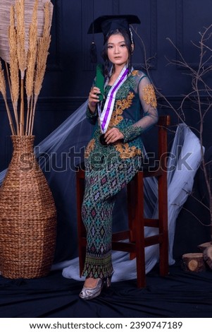beautiful Asian woman graduating standing in front of a chair smiling thinly holding a diploma wearing a green kebaya and elegant exotic toga strap in an indoor photo studio with a black background