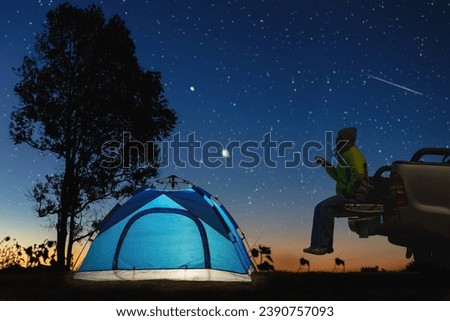 Male traveler sitting on car near blue camping tent looking at night sky with stars and Milky Way. Concept of travelling.