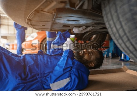 Mechanic in blue uniform lying down and working under car at auto service garage. Portrait of a happy mechanic man working on a car in an auto repair shop.